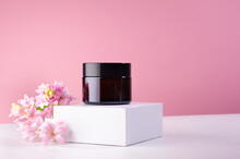 Cosmetic Jar For Cream Of Amber Glass On White Podium And Pink Wall With Fresh Spring Flowers. Template For Branding Identity For Cosmetics Produce.