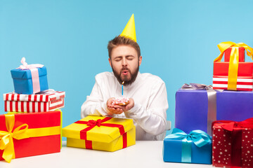 Funny bearded man in party cone making a wish closing eyes and blowing out candle on cake in his hands, celebrating birthday with many gifts at work. Indoor studio shot isolated on blue background