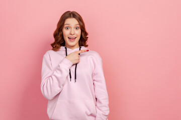 Poster - Portrait of surprised young female with curly hair in hoodie looking at camera with happy amazed expression, pointing finger right at copy space. Indoor studio shot isolated on pink background