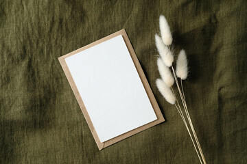 white invitation card mockup and dried flowers on green fabric top view. vintage wedding invite card