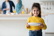 © Prostock-studio - Little Helper. Cute Arab Girl Holding Plates In Kitchen, Smiling At Camera