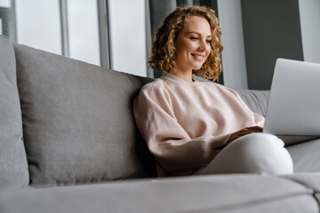 Wall Mural - White blonde woman smiling and using laptop while sitting on couch