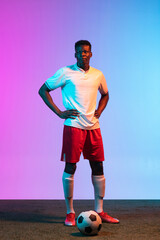 Wall Mural - Full -length portrait of African professional football player standing isolated on gradient blue pink background in neon light.