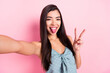Photo of funky happy nice young woman stick out tongue make selfie show v-sign isolated on pink color background