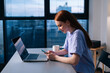 © dikushin - Side view of cheerful young woman using mobile phone sitting at desk with laptop near window with cityscape evening at late. Attractive redhead lady working from home office looking at screen of cell.