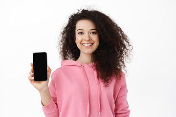 Portrait of brunette curly girl showing application on phone, smartphone empty screen, smiling as recommending download app, standing against white background
