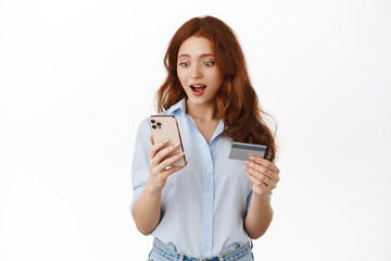 Wall Mural - Amazed redhead girl staring at smartphone, seeing discounts on shopping site, holding credit card, paying with mobile phone app, standing against white background