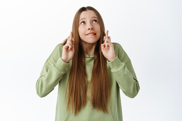 Wall Mural - Excited cute girl biting lip nervous, cross fingers and look up at copyspace, waiting for results with worried expression, anticipating news, praying, standing over white background