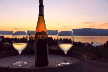 Dark sunset - romantic scene with the wine glasses and bottle of white wine on the wooden barrel by Okanagan Lake in Kelowna, BC