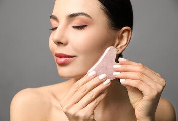 Poster - Beautiful young woman doing facial massage with gua sha tool on grey background, closeup