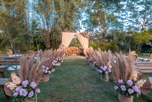 A Rustic Garden Wedding Setup, The Main Aisle Lined With Baskets With Pampas Leaves And Pastel Pink Roses. Trees In The Background.