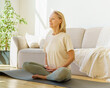 © Nata Bene - Peaceful senior woman in lotus position meditation with closed eyes at home while sitting on yoga mat on floor, full length. Calm elderly lady practicing meditation techniques and yoga indoors