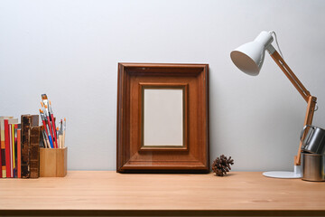 Stylish workplace with empty photo frame, lamp, pencil holder and book on wooden table.