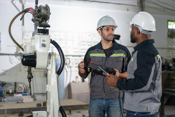 Wall Mural - Male engineers standing to discuss controlling a robot arm welding machine using a tablet and remote control cable connect to robot in an industrial factory.