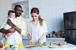 © LIGHTFIELD STUDIOS - happy african american man holding infant daughter near wife preparing breakfast