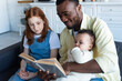 © LIGHTFIELD STUDIOS - smiling african american man reading book to interracial daughters at home