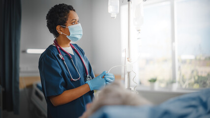 Wall Mural - Hospital Ward: Professional Black Head Nurse Wearing Face Mask Does Checkup of Patient's Vitals, Checking Heart Rate Computer, Intravenous or Iv Fluids Drip Bag. Caring Nurse Monitors Person Recovery