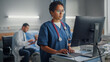 © Gorodenkoff - Hospital Ward: Professional Smiling Black Female Head Nurse or Doctor Wearing Stethoscope Uses Medical Computer. In the Background Patients in Beds Recovering Successfully after Sickness and Surgery