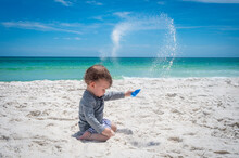Adorable Baby Beach Vacation Adventure Flipping White Sand Into The Air In Front Of Beautiful Blue Ocean With Plastic Shovel