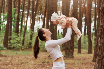 Wall Mural - Side view of young mother throws up baby in air while happy family playing together in forest, mom and little daughter, positive human emotions, feelings, joy.