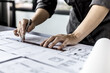 © kamiphotos - A close-up, an architect, a house designer holding a pen, pointing to a house plan to examine the design plan before discussing the details with the client. Interior design and decoration ideas.