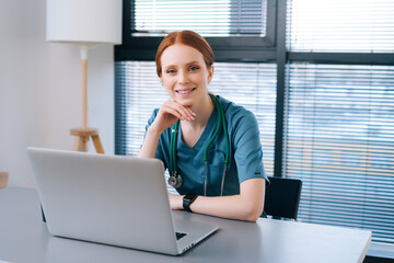 Portrait of attractive smiling young female doctor in blue green medical uniform sitting at desk with laptop on background of window in hospital office of medic clinic, looking at camera.