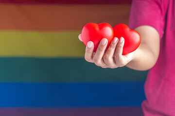 Hand holding two red hearts against the background of the rainbow flag (LGBT). Close-up photo. Love concept