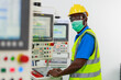 © butsaya33 - Portrait of African black man worker wearing protective face mask standing in front of control panel machine at the steel factory. Young African man in safety uniform with smiley face