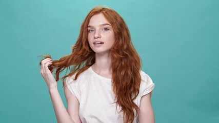 Poster - Happy shy ginger woman in t-shirt touching her hair and looking at the camera over turquoise background