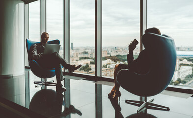 Silhouettes of two business coworkers relaxing in blue armchairs of a chillout office area in front of the window with a cityscape, with selective focus on a serious businessman with a laptop