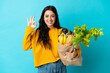 © luismolinero - Young woman holding a grocery shopping bag isolated on blue background showing ok sign with fingers