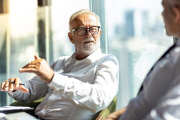 Picture of two business man discussing business and seem like an agreement is made. The young man is wearing white shirt with black tie. The older man is wearing black suit. They meet in hotel lobby.