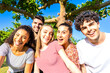 Group of five very happy friends having fun at the park posing looking at camera for a portrait. Gen z multiracial carefree young women and men together in a city park living nature in the summer sun