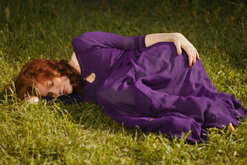 A beautiful young woman with red curly hair in a purple dress fell asleep in the garden on the grass.