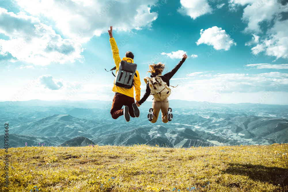 Hikers with backpacks jumping with arms up on top of a mountain - Couple of young happy travelers climbing the peak - Family, travel and adventure concept - obrazy, fototapety, plakaty 