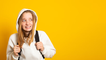 Wall Mural - Cute cool schoolgirl dressed white hoodie on yellow  background with backpack looking away. Banner copy space.