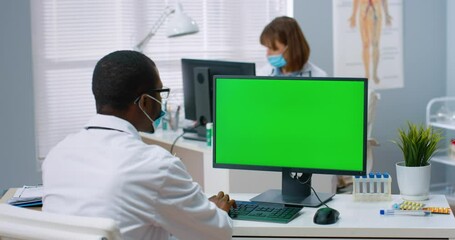 Young African-American doctor sits in hospital office and works at computer typing on keyboard. View from back. Chromakey.