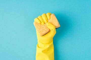 Top view photo of raised hand in yellow rubber glove clenching yellow scouring pad on isolated pastel blue background with copyspace