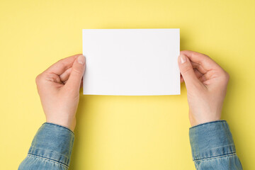 First person top view photo of female hands holding white paper card on isolated yellow background with blank space