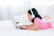 © Rakchanok - Adorable Asian girl concentrate online education class on laptop, kids lie down on bed watching game or social media connect to friends, smart school girl study e-learning using headphone technology