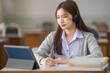 © EduLife Photos - Young Asian woman college student with wireless headphones watches and listens to the lecturer on tablet and writes on the notebook to study online by herself in classroom