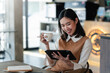 © amnaj - Happy Asian businesswoman relaxing holding a coffee cup and pen and watching tablet at a café.