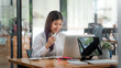 © amnaj - Smiling young Asian woman working on laptop and drinking coffee at office.