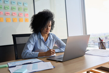 Young African American focused female ceo data analyst businesswoman sitting at desk with laptop writing notes near panoramic window in contemporary corporation office. Business technologies concept.