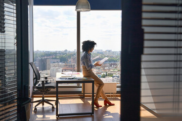 Wall Mural - Young successful female African American executive manager ceo sitting on desk reading financial report in modern office in high floor corporate building with panoramic urban megapolis city view.