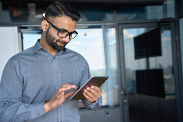 Young indian marketing business manager expert in glasses standing in modern corporate office using digital tablet device searching analyzing project results. Business technologies concept.