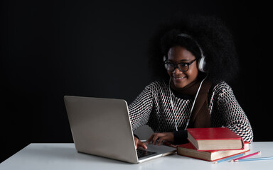 Portrait of African girl wear glasses and headphones smiling while online learning with laptop textbooks. Teenage student girl with sweater happy to study on black background. Copy space