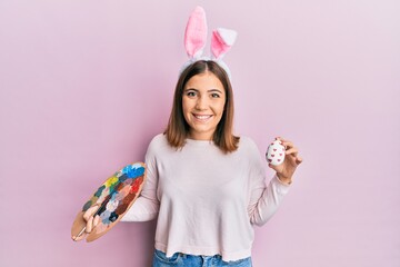 Poster - Young beautiful woman wearing cute easter bunny ears holding painting palette and egg smiling with a happy and cool smile on face. showing teeth.