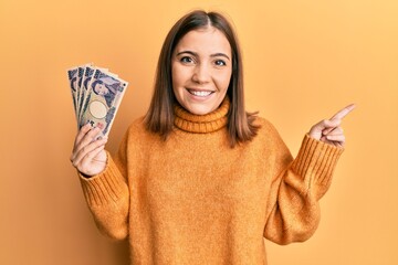 Wall Mural - Young woman holding 5000 japanese yen banknotes smiling happy pointing with hand and finger to the side