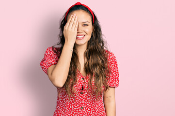 Young hispanic girl wearing casual clothes covering one eye with hand, confident smile on face and surprise emotion.